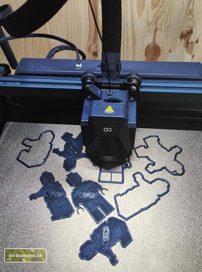 Blick auf's Druckbett eines 3D-Druckers, auf dem 4 verschiedene Cookie-Cutter und Stamps (neudeutsch für Keksausstecher und Stempel) aus schwarzem Filament gedruckt werden. Motive: Legomännchen in 4 verschiedenen Posen.