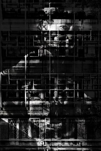 Black and white photograph, portrait format, multiple exposure. Self-portrait with architectural elements (Brutalist façade with many windows). Through a dark grid of windows dominated by the horizontal and vertical lines of the façade, the shadowy body and head of a woman are discernible. She has turned her back to the camera, but her head is turned so that it looks at the viewer as if through a window. She is reflecting herself into a mirror. The overall impression is gloomy, the subject's gaze critical and inquisitive; the architectural elements act like a prison in front of the body.