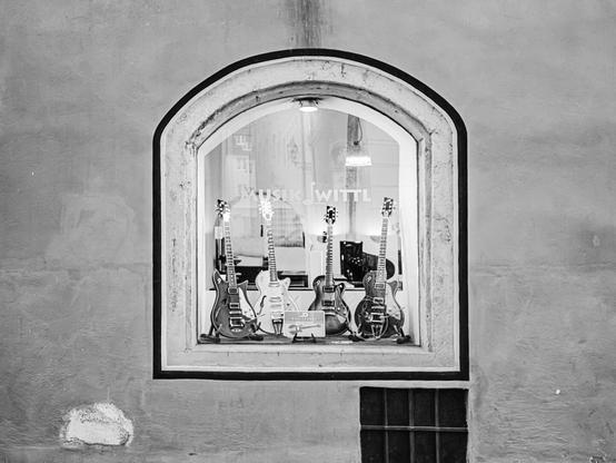 Black and white photo of a music store window displaying four electric guitars standing upright. The arched window is set into an aged stucco wall, with the text "MUSIK WITTL" partially visible on the glass.