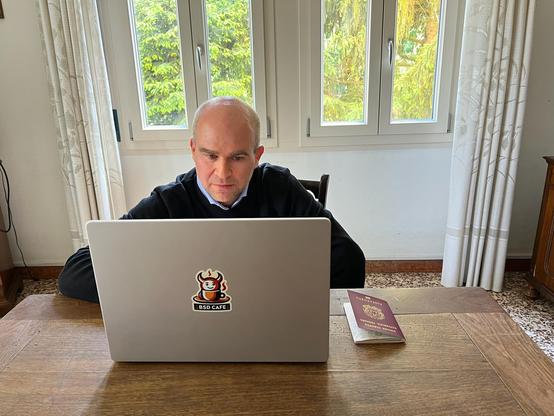 Man (me) sitting at a wooden table using a laptop with a prominent "BSD Cafe" sticker on the back. I'm focused on the screen,  applying for the Canadian eTA to attend BSDCan 2025. An open passport lies next to the laptop. Behind me, large windows let in natural light and show a lush, green outdoor scene.