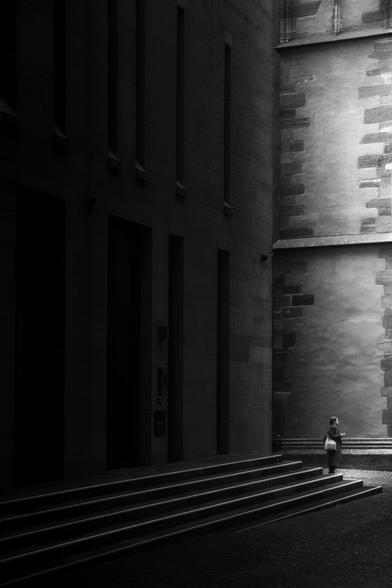 black and white. portrait format. the picture was taken in the densely built-up old town of frankurt. narrow alleys, some of which are very dark and hardly get any sunlight. in the picture a woman stands between two large, massive historical buildings. the left part of the picture is very gloomy. the woman stands in the right part of the picture. this part is brightly lit by the sun and forms a balancing contrast to the darkness. when i look at the picture i think of confidence, hope and faith.