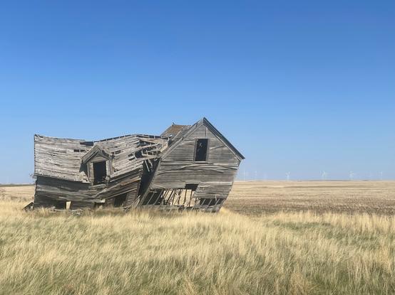 Old farmhouse falling down/leaning on the prairies with blue sky background and turbines