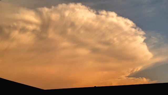 A towering thunderstorm cloud shading orange due to the setting sun as seen over a rooftop