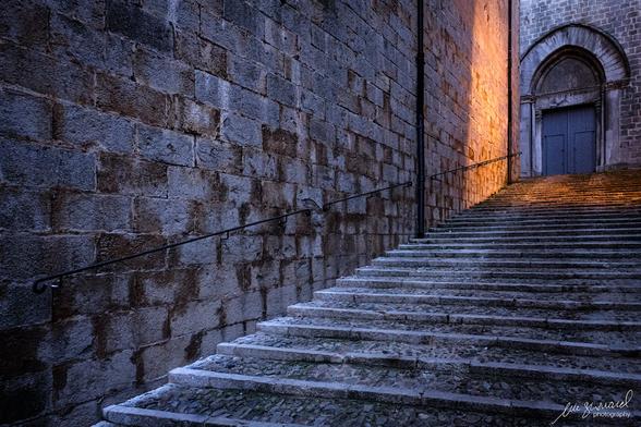 The setting sun hits an old stone wall and the top of a stone staircase that leads towards a door nestled in an archway in another stone wall in the ancient streets of old Girona, Spain, in 2019.