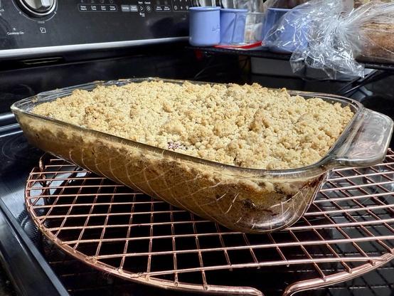 7x11 glass baking dish sitting on copper round cooling rack on black stove top. Inside the glass dish is a tan brown crumbly and lumpy looking coffee cake that smells divine. Can I resist eating it before it’s cool? Not sure I have the willpower.