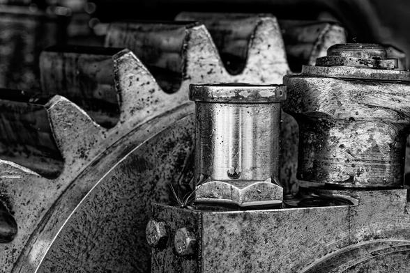 A close-up, black-and-white photograph of industrial machinery, featuring large metal gears and bolts. The gears appear worn and coated in grime, emphasizing their heavy use. The focus is on a cylindrical metal component with a hexagonal base, surrounded by intricate mechanical parts. The monochrome tones highlight the rugged textures and sturdy construction of the machinery.