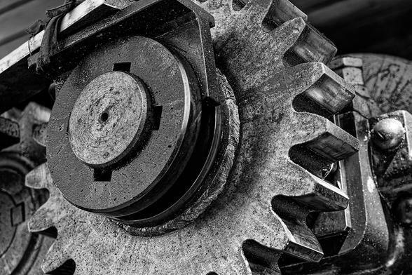 A close-up, black-and-white photograph of a large industrial gear mechanism. The central focus is a metallic gear with prominent, well-defined teeth, showing visible wear and scratches. Attached to the gear is a smaller circular component, appearing to be part of the machinery's inner workings. The monochrome palette emphasizes the rugged textures and intricate details of the metal surfaces, highlighting the craftsmanship and engineering involved in its construction.