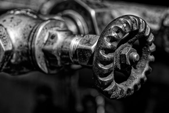 A close-up view of a metal valve handle, circular in shape with a textured, gear-like surface. The metal appears worn and slightly corroded, suggesting long-term use. The background is blurred, drawing attention to the intricate details of the valve handle and adjacent metal components. The aged condition and industrial aesthetic of the image hint at its role within a mechanical or plumbing system.