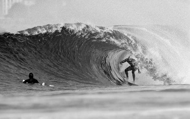 A monochrome shot of a surfer riding the barrel, as seen from a distance.