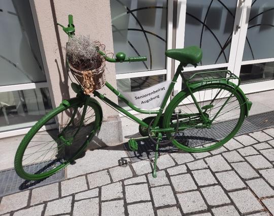 Vollständig grün angemaltes Fahrrad mit im Rahmen eingespanntem Label "Seniorenbeirat Augsburg" und Fahrradkorb vornedran.