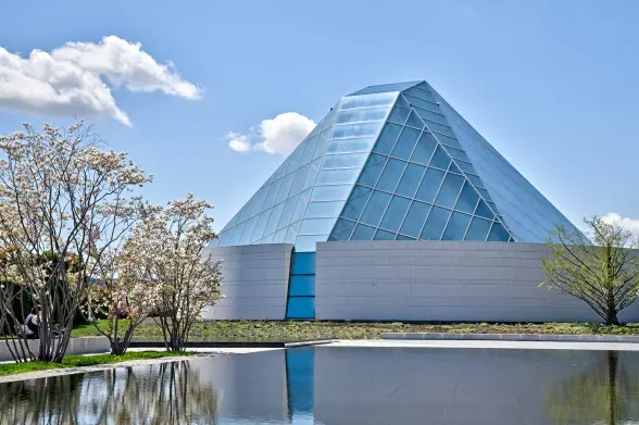 A bright daylight photograph of the Aga Khan Ismaili Centre in Toronto, featuring its distinctive glass pyramid structure. The pyramid has a blue-tinted geometric grid of glass panels rising from a white stone base. The modern building is reflected in a still reflecting pool in the foreground. Blooming trees (likely dogwood or cherry) frame the left side of the image, and the scene is set against a vivid blue sky with scattered white clouds. The contemporary Islamic-inspired architecture creates a striking contrast with the natural elements of water and flowering trees in the landscaped grounds.