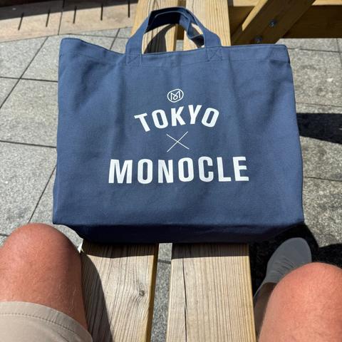 Close up of a navy blue canvas tote bag, which reads “Tokyo x Monocle” in white text. It’s resting on a bench in front of me and my knees are just visible poking out of my shorts - sorry! lol