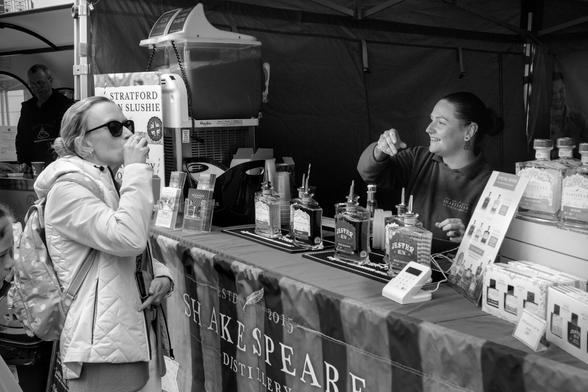 A woman stood at the counter of a gin stand. She is knocking back a drink. It's not visible, but the context of standing at the gin stand suggests it's a sample of the bottles being sold. On the counter are bottles of gin and rum on display. The other side of the counter a girl is enthusiastically talking, presumably about the gin she is selling. She is holding her hand up as if to knock back a shot of the drink. She is smiling, maybe laughing as she talks.