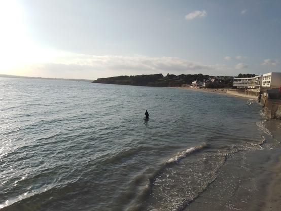 Photo de la plage des Sables Blancs à Concarneau, prise en début de soirée mi-mai. La marée est haute et on ne voit presque plus le sable. On aperçoit un pêcheur à pied dans l'eau, au centre de la photo. Au fond, après la plage, la côte devient plus abrupte et est très boisée.