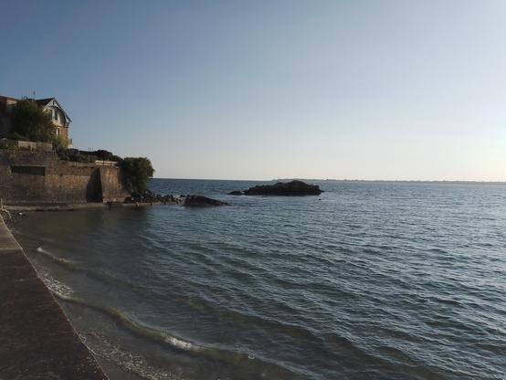 Photo prise depuis la plage des Sables Blancs à Concarneau. La mer est calme et haute. On aperçois une maison donnant sur la mer et un rocher qui émerge encore de l'eau. Au fond, on aperçoit la pointe de Beg Meil.