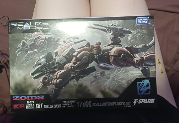 A photograph of the box art of the RMZ Hell Cat model kit, resting on Beatrix's thighs. The box depicts a few examples of the Zoid in question, nominally a panther but generally just a small feline with an interesting use of the standard clamshell cockpit with a little kitty-eared "helmet" over it. They're running in a jungle, the environment most associated with the Zoid.

Beatrix's right thigh has a gay daisy band-aid on it for v. homosexual HRT reasons.