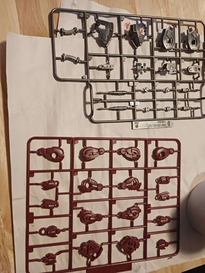 A photograph of two lightly-detailed Zoid sprues drying on the crafting table. There's a gunmetal sprue of various parts with four similar pieces which have had a small shaft painted silver, and a red sprue with piston halves on leg pieces painted gold, and some screws on other leg pieces painted silver.