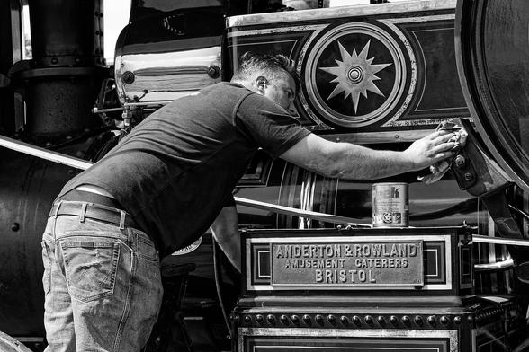 A black-and-white photo shows a person cleaning a vintage steam engine. The individual is dressed in jeans and a dark shirt, leaning forward with a cloth in hand, carefully polishing the engine's ornate details. The steam engine has a decorative star emblem and a plaque that reads "ANDERTON & ROWLAND AMUSEMENT CATERERS BRISTOL." A can of Peek polish sits on a ledge near the plaque, emphasizing the restoration effort.