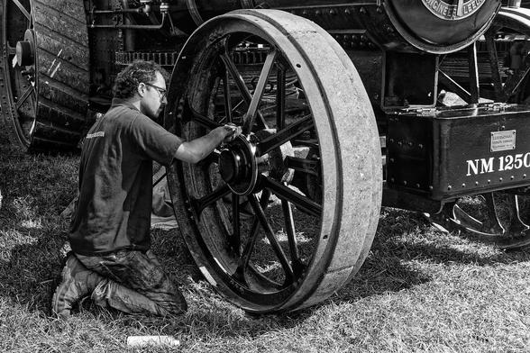 A black-and-white photograph of a person kneeling on the grass, working on the large wheel of a vintage steam engine. The wheel has a solid rubber tire and multiple spokes. The steam engine features exposed gears and intricate mechanical components. There are markings on the engine, including a label that reads "NM 1250" and a plaque that says "Teledyne Engineering Ltd." The person is dressed casually in a shirt and pants, appearing focused on maintenance or repair work. The outdoor setting, with grass visible beneath the engine, adds to the historical, industrial atmosphere.