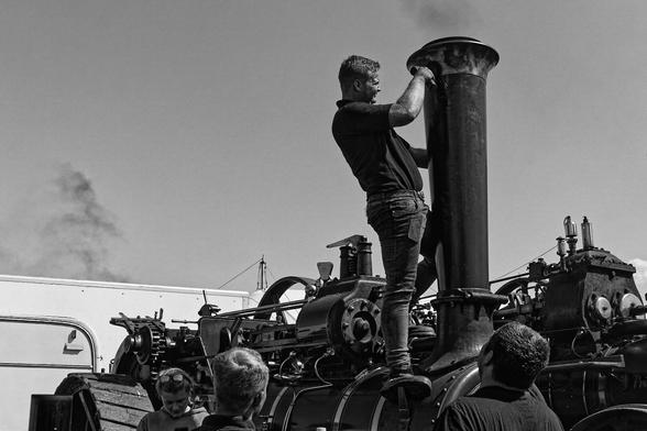 A black-and-white photograph captures a person standing on a large piece of machinery, possibly a steam engine or an old industrial machine. The individual holds onto a tall, cylindrical structure that appears to be a smokestack or chimney. Several other people stand nearby, looking up at the person. The machinery is filled with gears, pipes, and mechanical components, emphasizing its industrial nature. In the background, a building is visible, with smoke rising into the sky, suggesting the machinery may be in operation or undergoing maintenance.