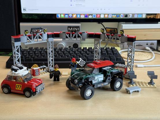 A LEGO Speed Champions set on a desk, featuring a red classic Mini Cooper in a rally configuration with rally lights and spare tires mounted on the roof of the car and tagged with number 177, a green John Cooper Works MINI Cooper in a rally configuration with a rally suspension and large wheels, a mechanic’s jack and wheeled bed, a trophy, and a woman holding a walkie-talkie and a man holding a checkered flag and a camera. The cars are in front of a combination finish line podium and mechanic’s bay that has LEGO screens showing highlights of each of the cars racing.