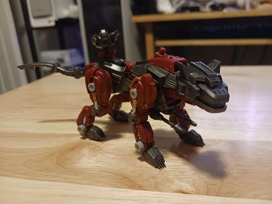 A photograph of the assembled helcat. It's a very small model, only about six inches tip to tail, in a mixture of gunmetal grey and a darkish red (though not quite burgundy). The legs each have a piston and screw which are painted in their traditional gold and silver, and the forelegs have copper tubing on the back of the shoulder as well. It has a shock cannon which looks oversized for it on the underside, and a basic two-barrel gun on the back. There's an unpopulated 3mm peg on its back between its front legs, an odd holdover from the original '80s model kits which the RMZ line have mostly thus far ignored in other cases.