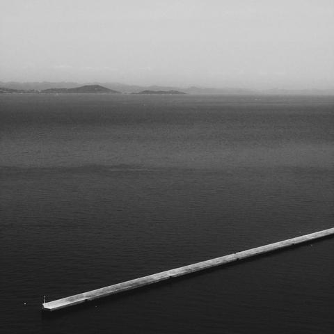 The image is a black and white photograph showing a body of water with a long, narrow pier extending into it. In the background, there are distant mountains or hills visible across the water. The scene appears calm and serene.
