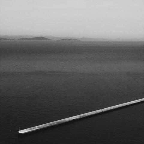 The image is a black and white photograph showing a body of water with a long, narrow pier extending into it. In the background, there are distant mountains or hills visible across the water. The scene appears calm and serene.