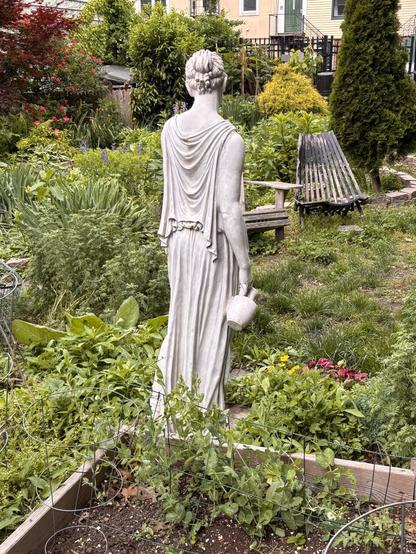 A plaster resin statue of a woman in a toga holding an urn in her right hand. The view is from behind, and her pose is relaxed, with her head slightly tilted. Around her are community garden plots and in the background some outdoor furniture.
