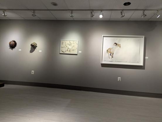 A wall of a gallery lit by spotlight. There are two animal masks on the left, and on the right two images of goats made out of dried flowers