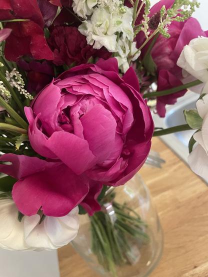 A close-up of a floral arrangement featuring a prominent pink peony, surrounded by white and burgundy flowers and greenery, placed in a glass vase.