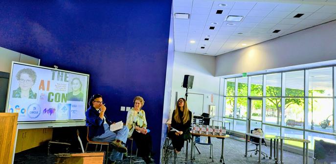 large open room with blue wall and three people sitting on stools: Alex Hanna, Emily M. Bender and Rhonda Holberton in discussion about the book, The AI Con.
