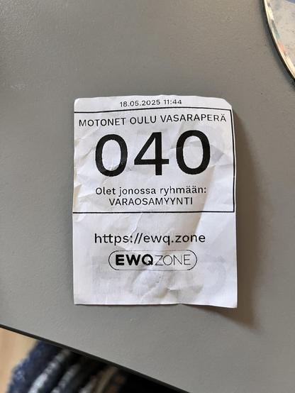 Mustavalkoinen jonotuslipuke: "18.05.202 11:44 Motonet Oulu Vasaraperä 040 Olet jonossa ryhmään: Varaosamyynti https://ewq.zone EWQZone" EWQZone-