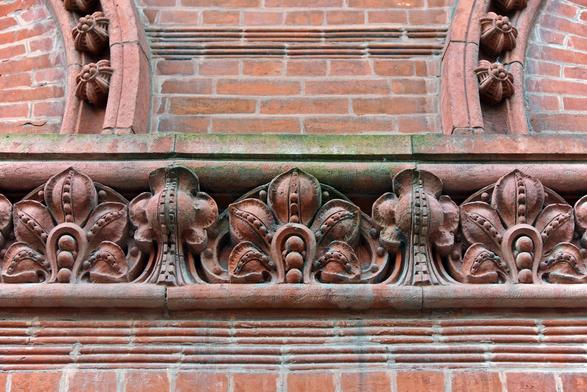 Color close-up of a band of brownish-red terra cotta ornament - executed in the Romanesque Revival style - set into a brick wall, just beneath a pair of arches