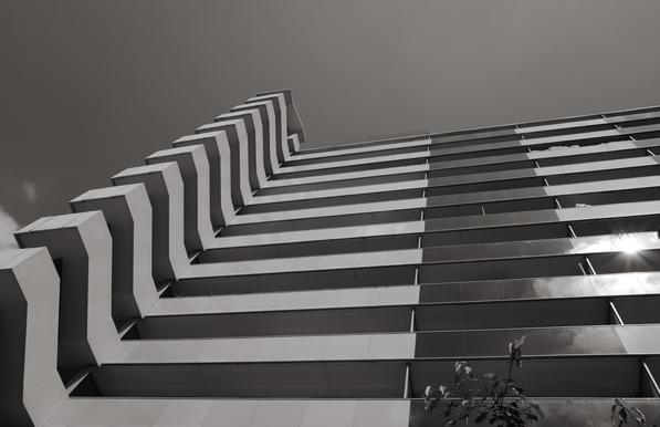 Un immeuble pris en contre plongée avec le ciel dans la partie supérieure et les différents balcons alignés avec l’escalier extérieur pour produire une continuité des lignes, tout en géométrie.
La photo est noir et blanc
