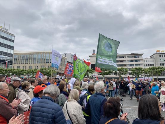 Links die Menschen der Demo, darüber wehen Fahnen: Verkehrswende ist Klimaschutz (VCD); A5 zehnspurig nicht mit uns; Vegans for future; Grandparents for future FFM: Wir haben die Erde nur geliehen)
Am rechten Bildrand weitere Menschen, eine Frau spielt Akkordeon.
Im Hintergrund Platanen und die Häuser neben der Konsti.