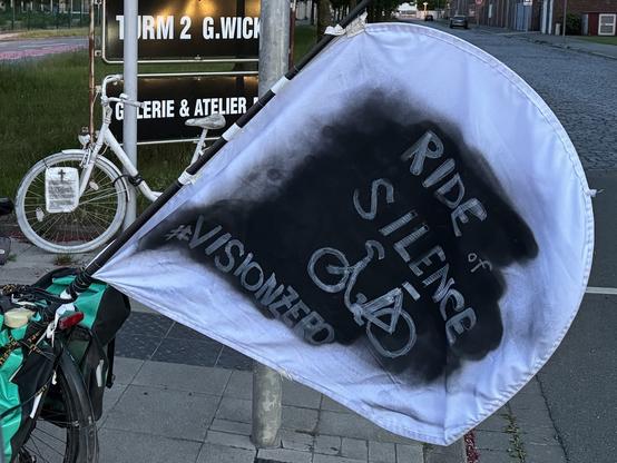 Foto. Eine Fahne aus weißem Stoff ist an einem Fahrrad befestigt. Ein Teil der Fahne ist mit schwarzer Farbe besprüht. Davon hebt sich in weißer Schrift der Text „Ride of Silence“ und die Silhouette eines Fahrrads ab. Im Hintergrund steht ein weißes Ghostbike.