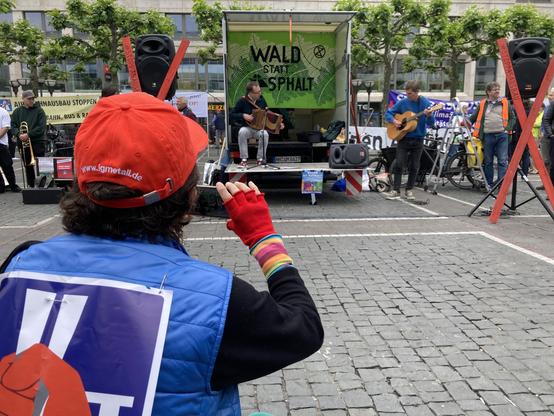 Im Vordergrund eine Frau mit roter Kappe, bunten Handschuhen und BI-Logo auf dem Rücken, die ein Foto von der LKW-Bühne macht. Auf und neben der Bühne spielt ein Duo Akkordeon und Gitarre. Hinter ihnen hängt ein grünes Banner von Wald statt Asphalt. Neben der Bühne stehen Lautsprecher, an denen rote Holzkreuze lehnen. Links der Bühne wartet ein Posaunist.
Im Hintergrund Platanen.
