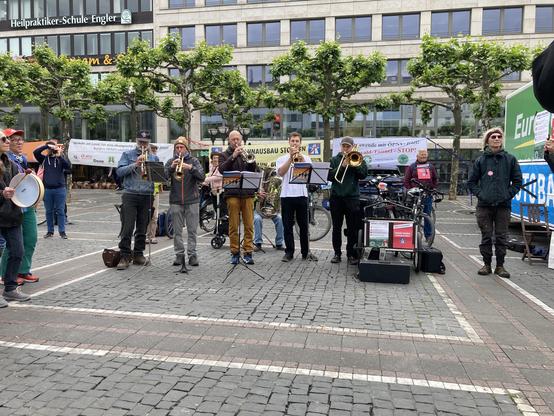 6 Bläser mit Trompeten, einer Posaune und einer Tuba stehen hinter Notenständern und spielen. Im Hintergrund Platanen und die Häuser neben der Konsti. Und Banner: Verkehrswendebündnis, Autobahnausbau Stoppen, Verkehrswende mit ÖPNV jetzt, Riederwald-Tunnel STOP!