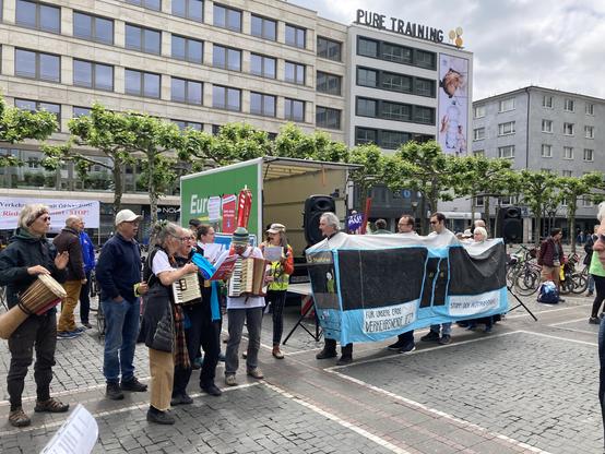 Blick Richtung LKW-Bühne. Links 7 Personen, die Akkordeon und Djembe spielen und singen, rechts 4 Personen, die gemeinsam in einem blauen Straßenbahn-Kostüm stecken. Die Straßenbahn ist ca 3 m lang und aus Stoff, der auf ein Gestell gespannt ist. Es schauen nur oben die Köpfe und unten die Füße raus. Auf der Straßenbahn steht: Für unsere Erde: Verkehrswende jetzt! Stopp den Autobahnbau!