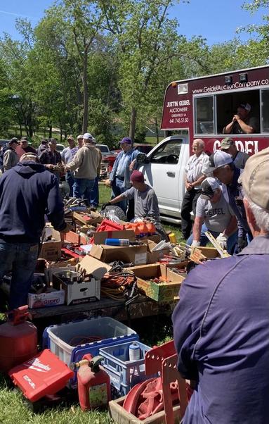 All manner of tools and shop items on tables and on the ground — part of the “lifetime accumulation” of an older man who recently died.  Men with baseball caps standing around. The auctioneer presides from an “office” on the back of a truck.