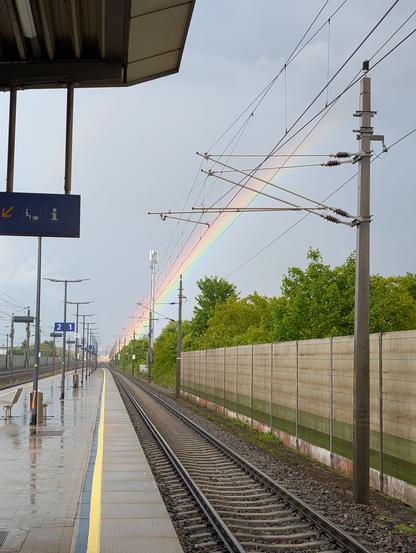 View along a train platform in the rain. A bright rainbow is visible behind the overhead catenary wire.