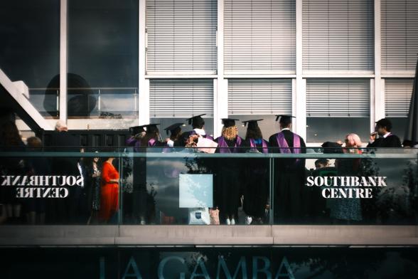 On the upper level outside the Royal Festival Hall on London's South Bank, a large number of graduates have gathered after the end of their graduation ceremony inside one of the theatres. They are drinking and chatting, comparing their future careers. They are wearing black gowns and mortar board hats, each with a maroon-coloured sash over their shoulders. We are looking at a row of seven of them, seen from behind, as they have their photo taken.