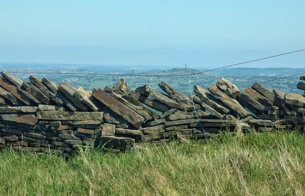 Grass, drystone wall, distant vista with tower topped salient hill emerging part way to the hazy horizon on a sunny day