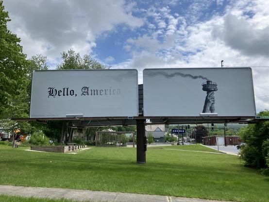 Two adjacent billboards: Left one has the text "Hello, America" in an old-fashioned (Gothic?) font, with the trailing letters of "America" gradually fading out. Right one has the hand and torch of the Statue of Liberty, with the flame burned out and smoke blowing into the left one. Art by Vincent Valdez.