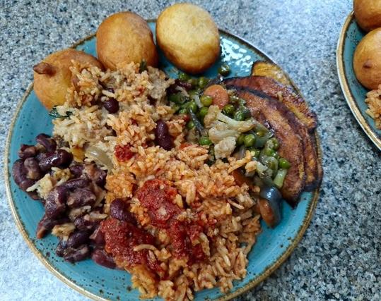 A shared serving of Mami Maggie's Recipes' Box of Everything lies on a brown-edged blue plate. Clockwise from the top: Three puff puffs, fried plantain, sautéed vegetables, jollof rice with tomato stew and scotch bonnet sauce condiments, sautéed beans and fried rice.