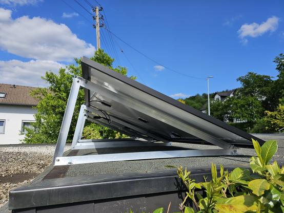 Das Bild zeigt ein Solarpanel auf dem Dach eines Gebäudes. Das Solarpanel ist an einem silbernen Metallrahmen montiert, der es ermöglicht, das Panel zu schwenken. Das Panel ist leicht geneigt und zeigt die Rückseite mit den elektrischen Verbindungen. Im Hintergrund sind Bäume und ein klarer blauer Himmel mit ein paar weißen Wolken zu sehen. Ein Strommast mit Leitungen ist ebenfalls sichtbar. Das Dach ist mit einem dunklen Material bedeckt, und vor dem Solarpanel wachsen grüne Pflanzen. Die Umgebung wirkt ruhig und sonnig, was die Effizienz des Solarpanels unterstreicht.