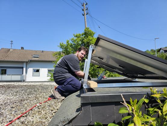 Ein Mann in einem dunkelgrauen T-Shirt und dunkelblauen Hosen sitzt auf einem Dach und installiert eine Solarpaneeleinheit. Er hält die Paneele mit einer Hand fest, während er mit der anderen Hand an der Montage arbeitet. Die Solarpaneele sind an einem Metallrahmen befestigt, der auf dem Dach platziert ist. Ein roter Kabelzug führt von der Paneele zum Dachrand. Im Hintergrund ist ein weißes Haus mit einem braunen Dach und einiger Vegetation zu sehen. Der Himmel ist klar und blau, was auf einen sonnigen Tag hinweist.