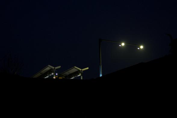 Night photo of the end of a couple of train platforms, flattened Y-shapes, taken from some way below. They're illuminated, perhaps, by a couple of bright lamps on the long arm of a lamppost to their right.