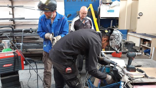 Poste soudure MIG et 3 hommes dans un atelier de travail du métal, au chevet d'un vélo de ville MBK bleu.